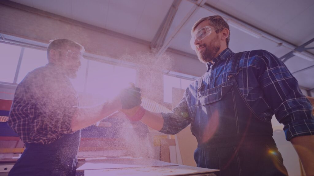 Two male factory workers in protective gear shaking hands in a workshop, surrounded by sawdust and sunlight