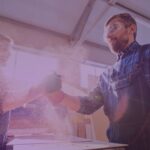 Two male factory workers in protective gear shaking hands in a workshop, surrounded by sawdust and sunlight
