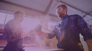 Two male factory workers in protective gear shaking hands in a workshop, surrounded by sawdust and sunlight